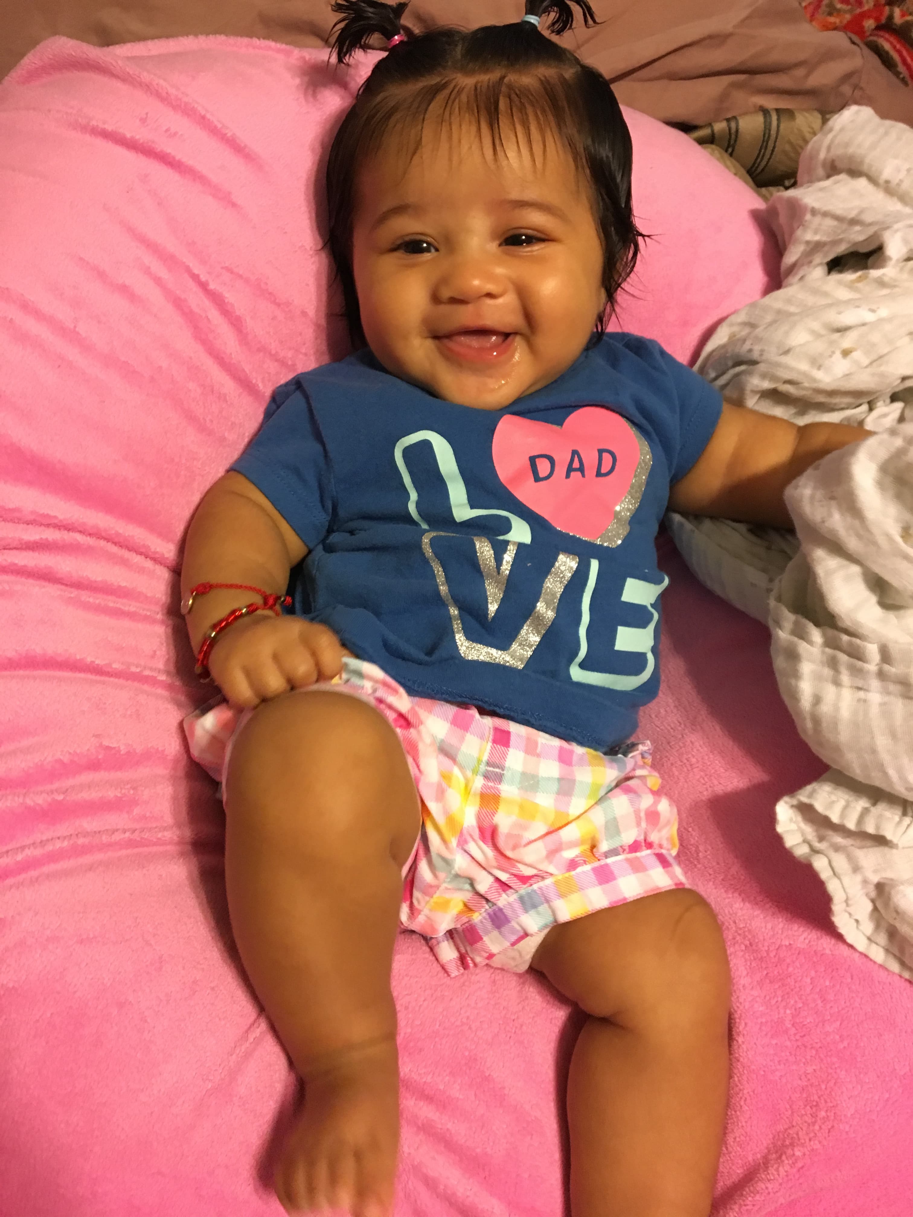 Jamie's granddaughter smiling, wearing a navy LOVE t-shirt with a heart that says DAD, on a pink blanket